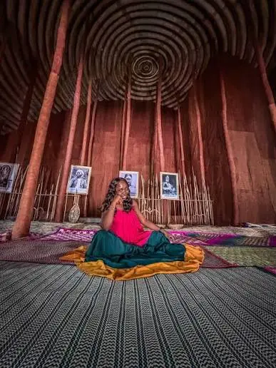 A woman in a vibrant dress sits on a patterned mat inside a building with tall red columns and a circular ceiling, reminiscent of places you might visit during the best safaris in Uganda. Black-and-white portraits are displayed on the wall behind her, adding to the cultural ambience.