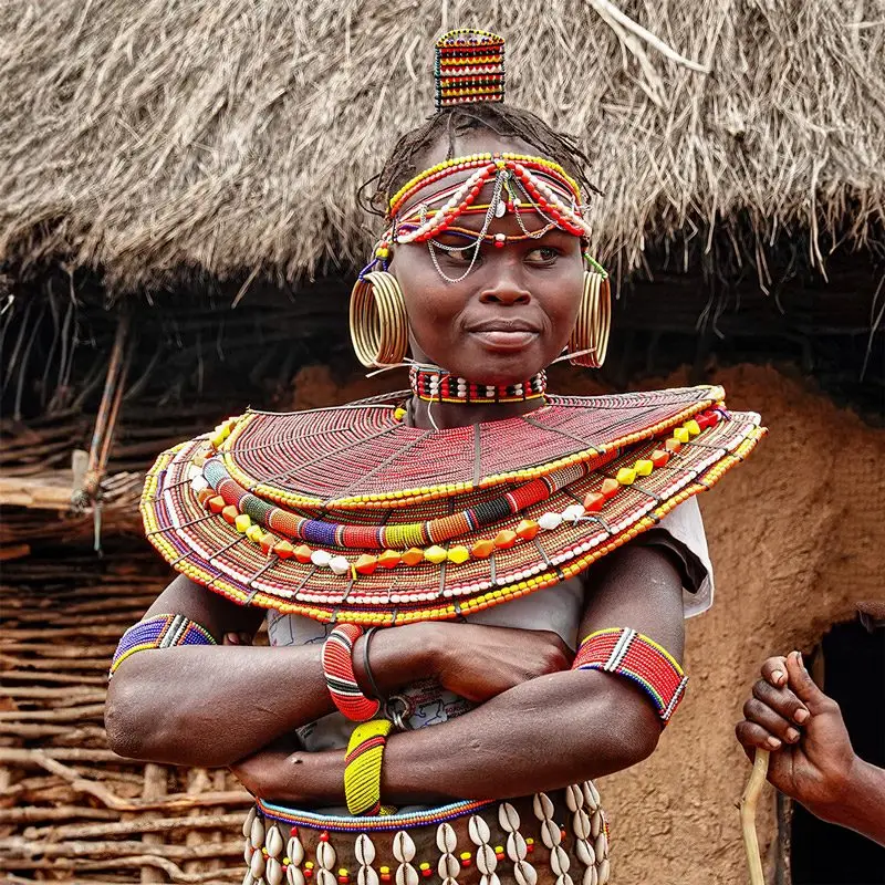A woman in traditional Maasai attire with colourful beaded jewellery, large earrings, and brass bracelets stands with arms crossed in front of a thatched-roof hut—a vivid image experienced on our Cultural Tours & Tribes Encounter Packages.