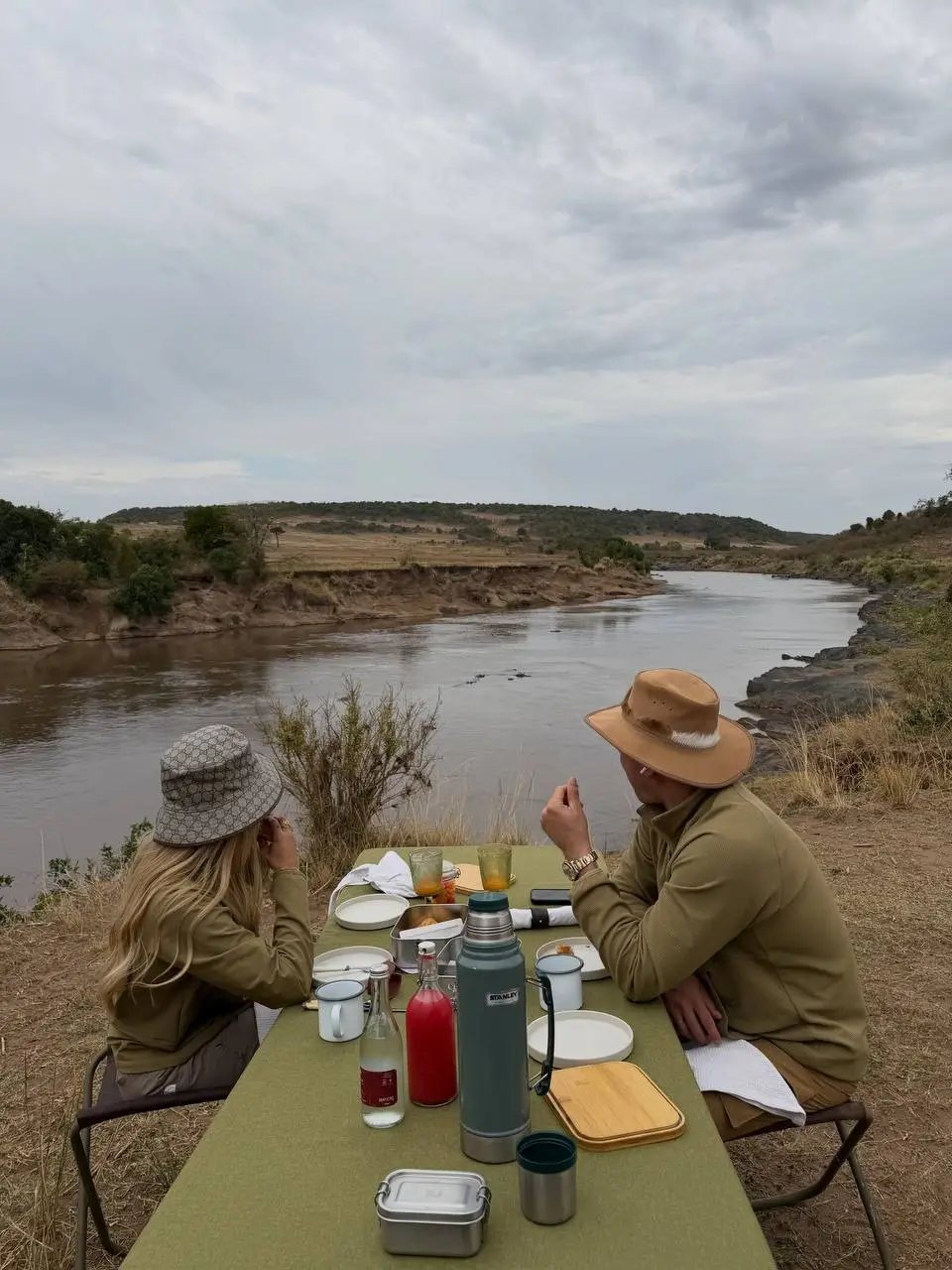 Two people in hats sit at a table set for a meal by a river, surrounded by savannah landscape under a cloudy sky—an idyllic pause during their wildlife adventures. Plates, cups, and drinks rest on the green tablecloth.